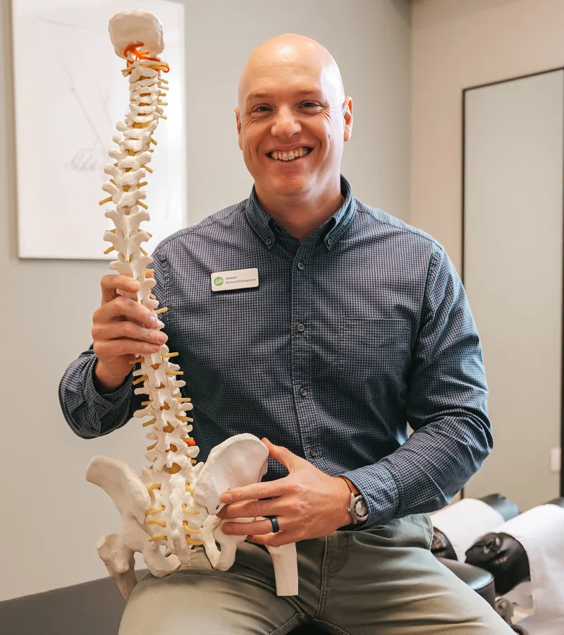 Smiling chiropractor holding an anatomical spine model in a treatment room